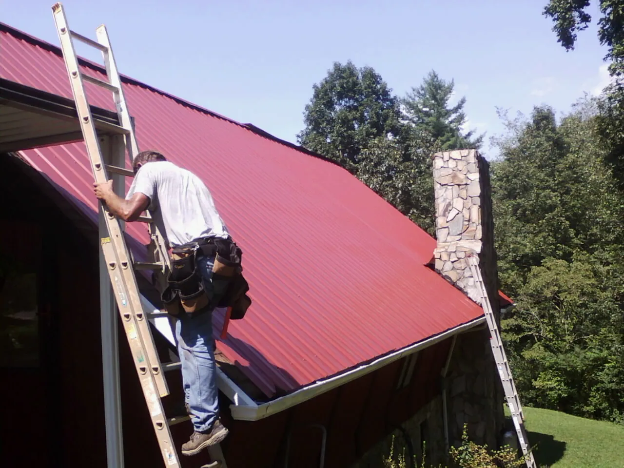 Roofer on a ladder beside a red metal roof and stone chimney — Soffit & Fascia Repair by Roofing Brothers in Gunlock
